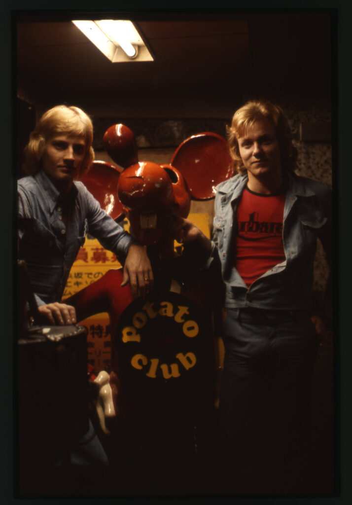 Två unga män på var sida en röd staty av en mus eller råtta, med skylten "Potato club".