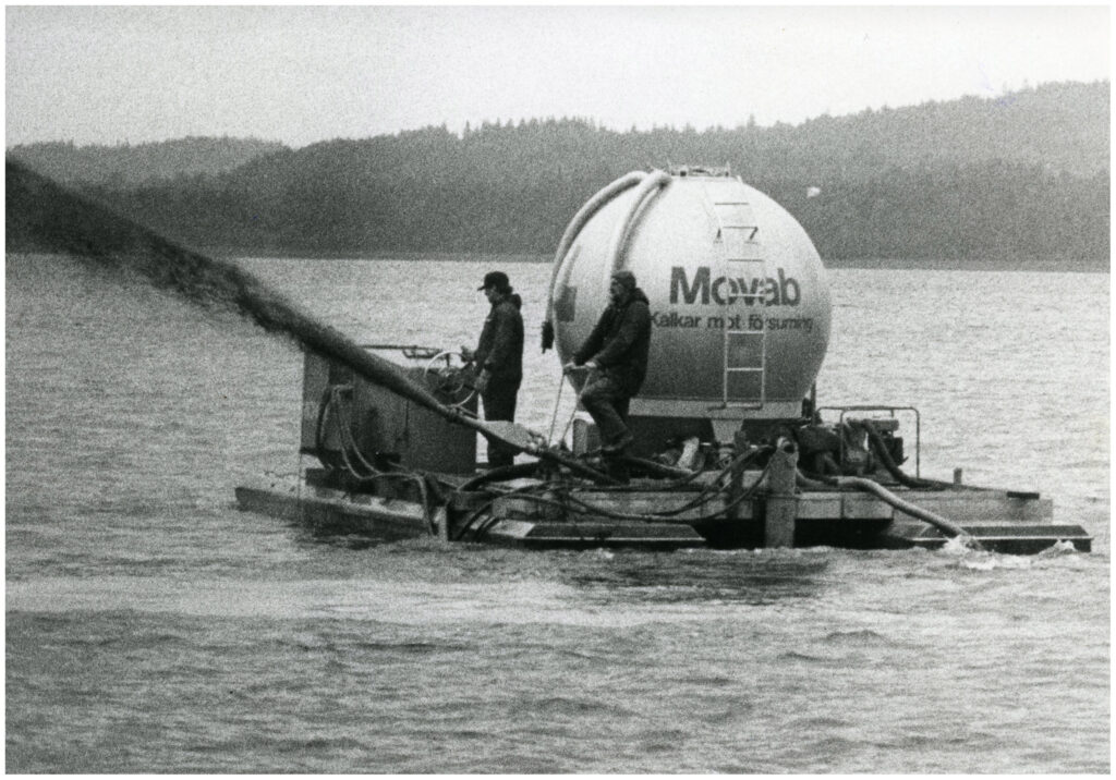 Två män på ett vattengående fordon som sprutar kalk med en slang ur en stor rund tank.