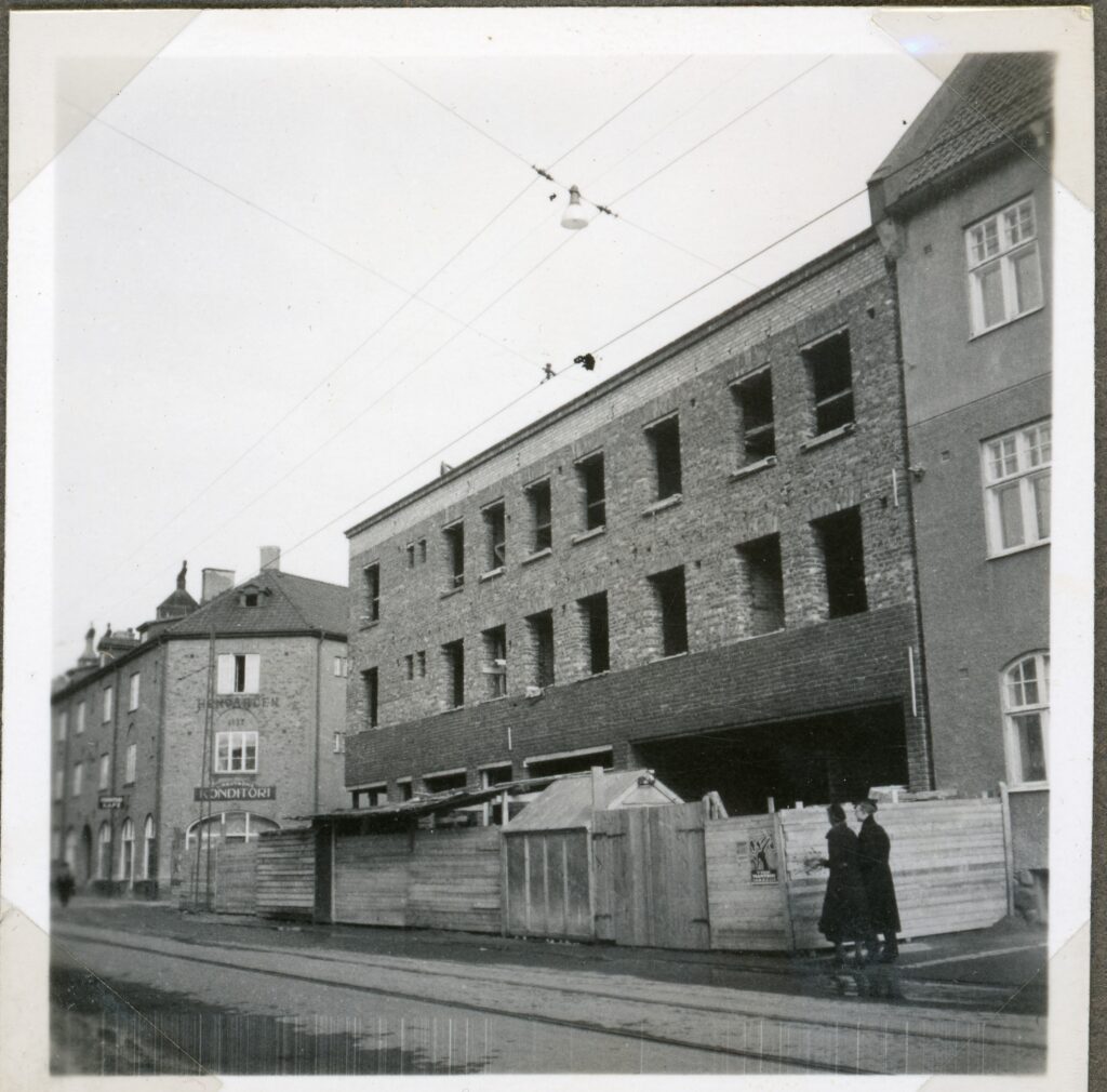 Äldre foto av byggnad under konstruktion med två damer i förgrunden och Lindströms Konditori i bakgrunden. 